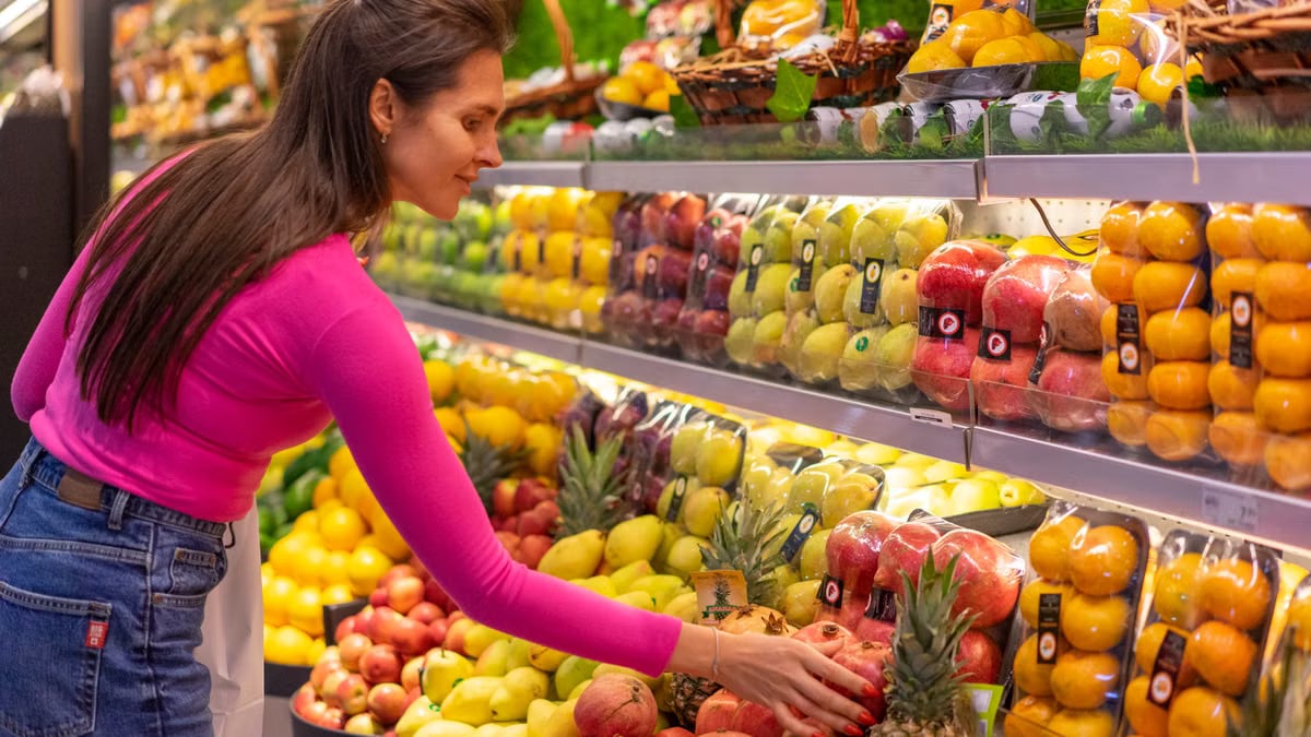 A woman in a bright pink top selecting fresh fruits in a grocery store produce aisle.